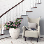 Beige armchair with a gray pillow and book beside a large potted plant in an Abigails White Ceramic Planter - Large, adorned with pink flowers, situated near a staircase with white spindles.