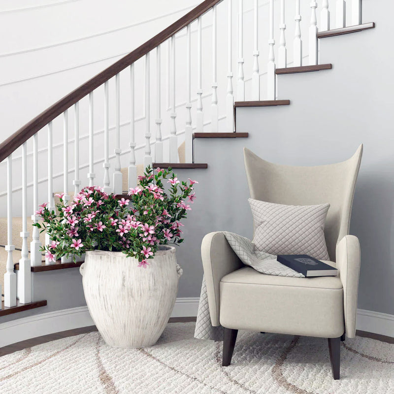 Beige armchair with a gray pillow and book beside a large potted plant in an Abigails White Ceramic Planter - Large, adorned with pink flowers, situated near a staircase with white spindles.