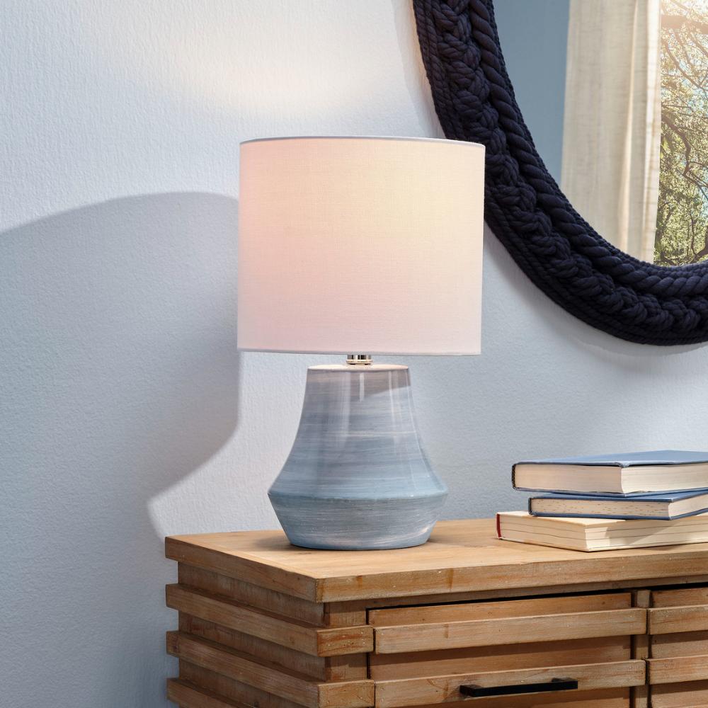 Table lamp with a textured base on a wooden surface, with a mirror and books in the background.