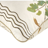 Close-up of a decorative throw pillow corner showing a cream fabric base with embroidered wavy dark brown lines, green leaves, and small brown bird motifs, finished with a gold-toned piped edge.