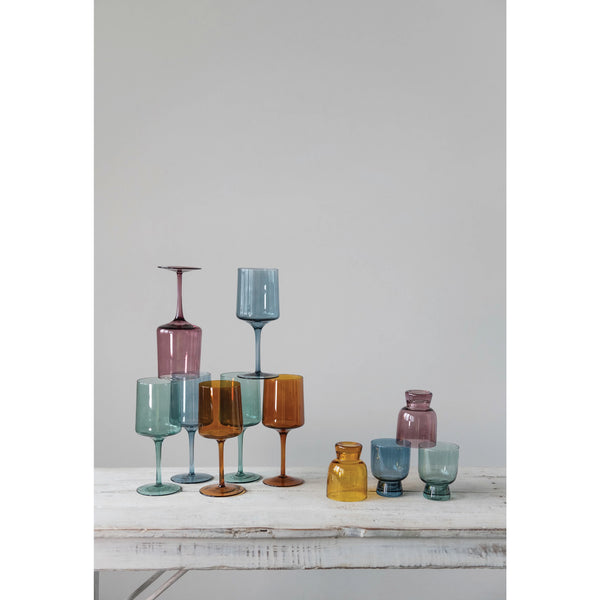 A set of four Colored Stemmed Wine Glasses by Creative Co-Op, boasting eye-catching hues and a 14 oz. capacity, is elegantly arranged on a white wooden table against a simple backdrop.