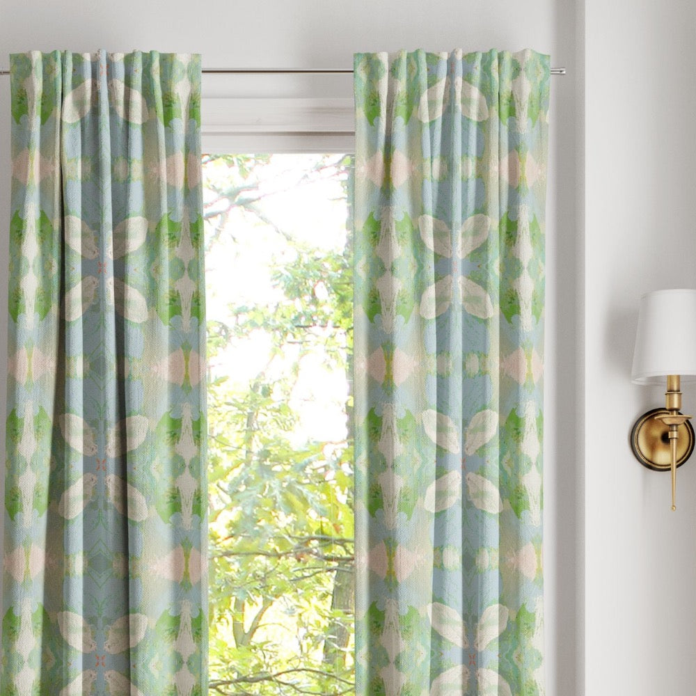 Laura Park Designs’ Elephant Falls Curtain Panels in light blue and green frame a window with leafy trees outside.