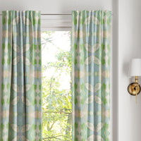 Laura Park Designs’ Elephant Falls Curtain Panels in light blue and green frame a window with leafy trees outside.