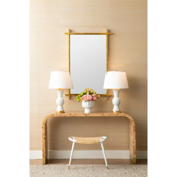 A styled interior scene showing a console table against a beige wall, with a rectangular mirror above it, two white table lamps, a small flower arrangement, and a stool in front.