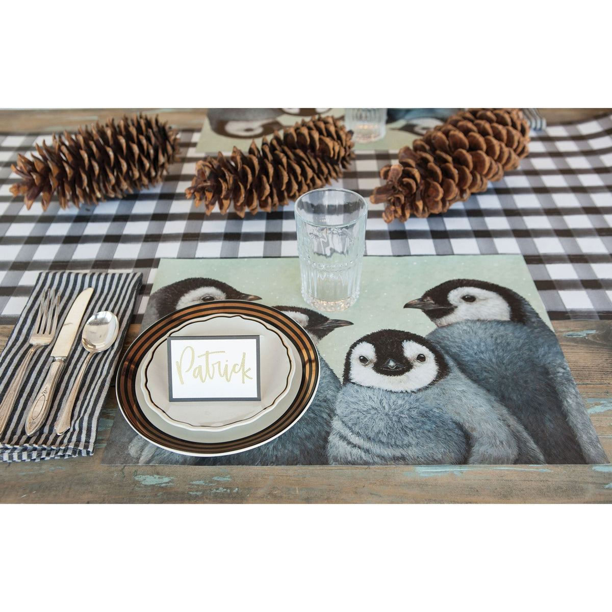 A delightful holiday party tablescape displays BSEID's Winter Penguin Paper Placemats, complemented by pinecones, cutlery, and a card with "Patrick" on the plate.