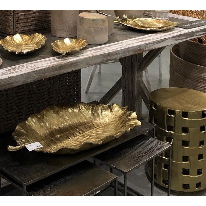 Alegra Leaf Trays by Napa Home & Garden, a set of two gold, leaf-shaped handcrafted aluminum trays, beautifully presented on display with rustic decor and a woven basket.