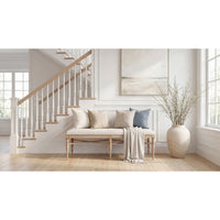 A styled interior scene showing a light wood bench with cream upholstery and pillows, a draped throw blanket, a staircase in the background, and a large beige vase with dried branches on a light wood floor.