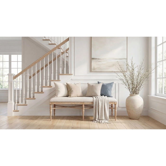A styled interior scene showing a light wood bench with cream upholstery and pillows, a draped throw blanket, a staircase in the background, and a large beige vase with dried branches on a light wood floor.