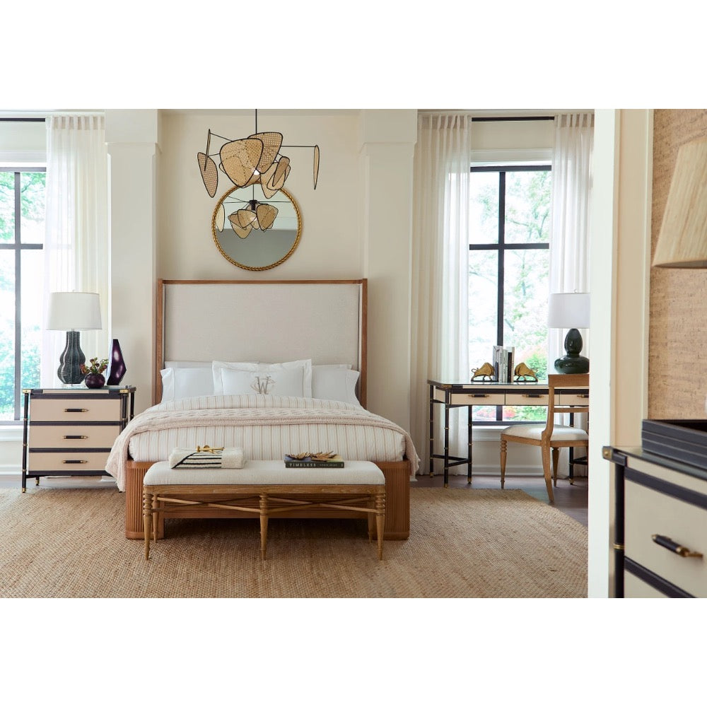 A styled bedroom interior featuring a bed with a light beige upholstered headboard, striped bedding, a matching bench at the foot of the bed, bedside tables with lamps, a desk area, and a decorative wall mirror above the bed.