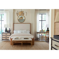 A styled bedroom interior featuring a bed with a light beige upholstered headboard, striped bedding, a matching bench at the foot of the bed, bedside tables with lamps, a desk area, and a decorative wall mirror above the bed.