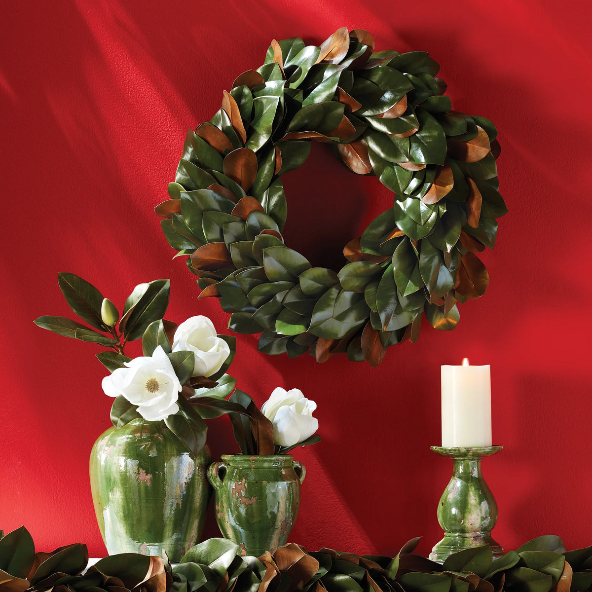 A Grand Magnolia Leaf Wreath by Napa Home & Garden adorns a red wall, complemented by green vases holding white flowers and a lit white candle on the table, creating a charming scene perfect for interior use.