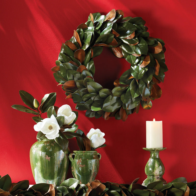 A Grand Magnolia Leaf Wreath by Napa Home & Garden adorns a red wall, complemented by green vases holding white flowers and a lit white candle on the table, creating a charming scene perfect for interior use.