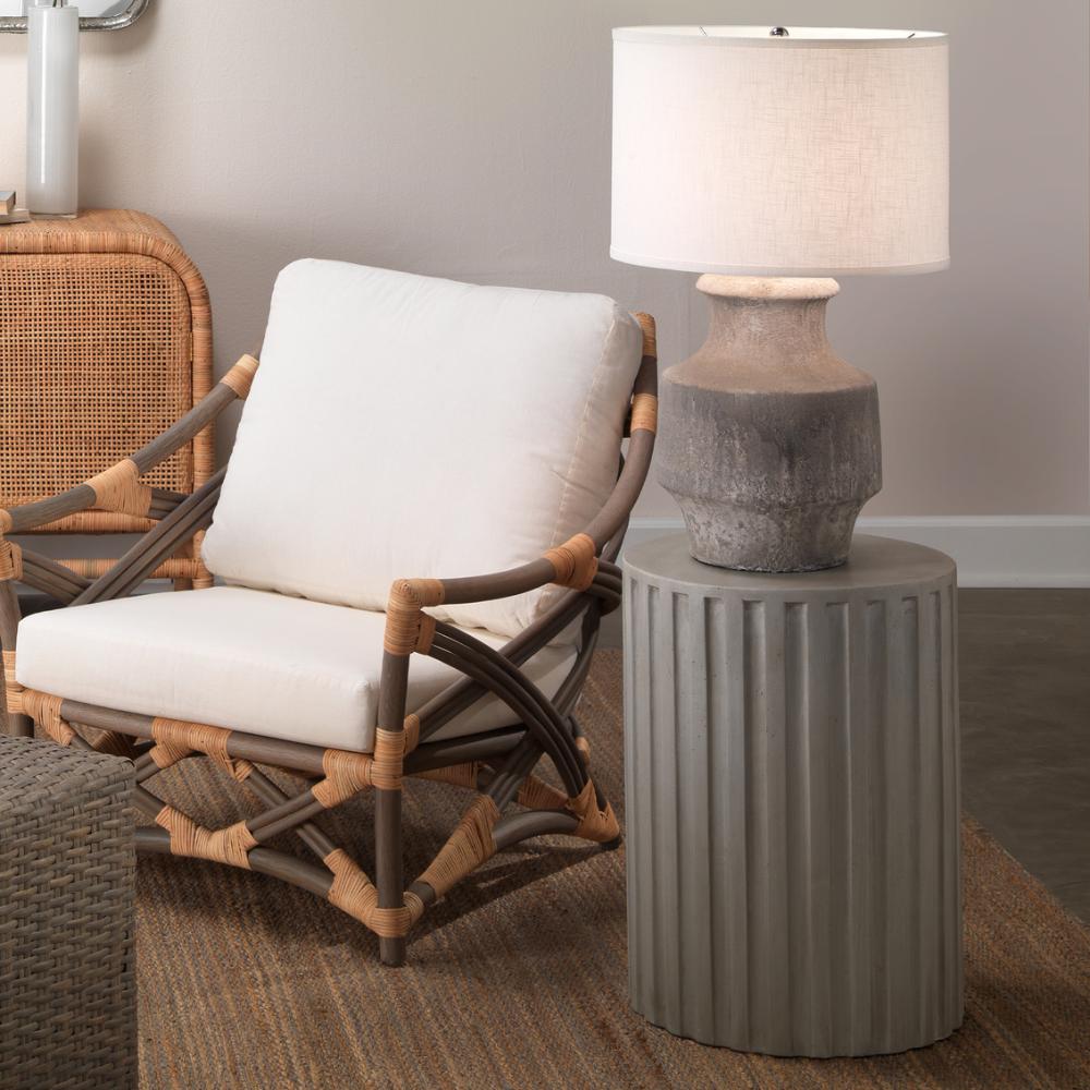Rattan chair with white cushions next to a gray side table with a lamp.