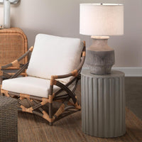 Rattan chair with white cushions next to a gray side table with a lamp.