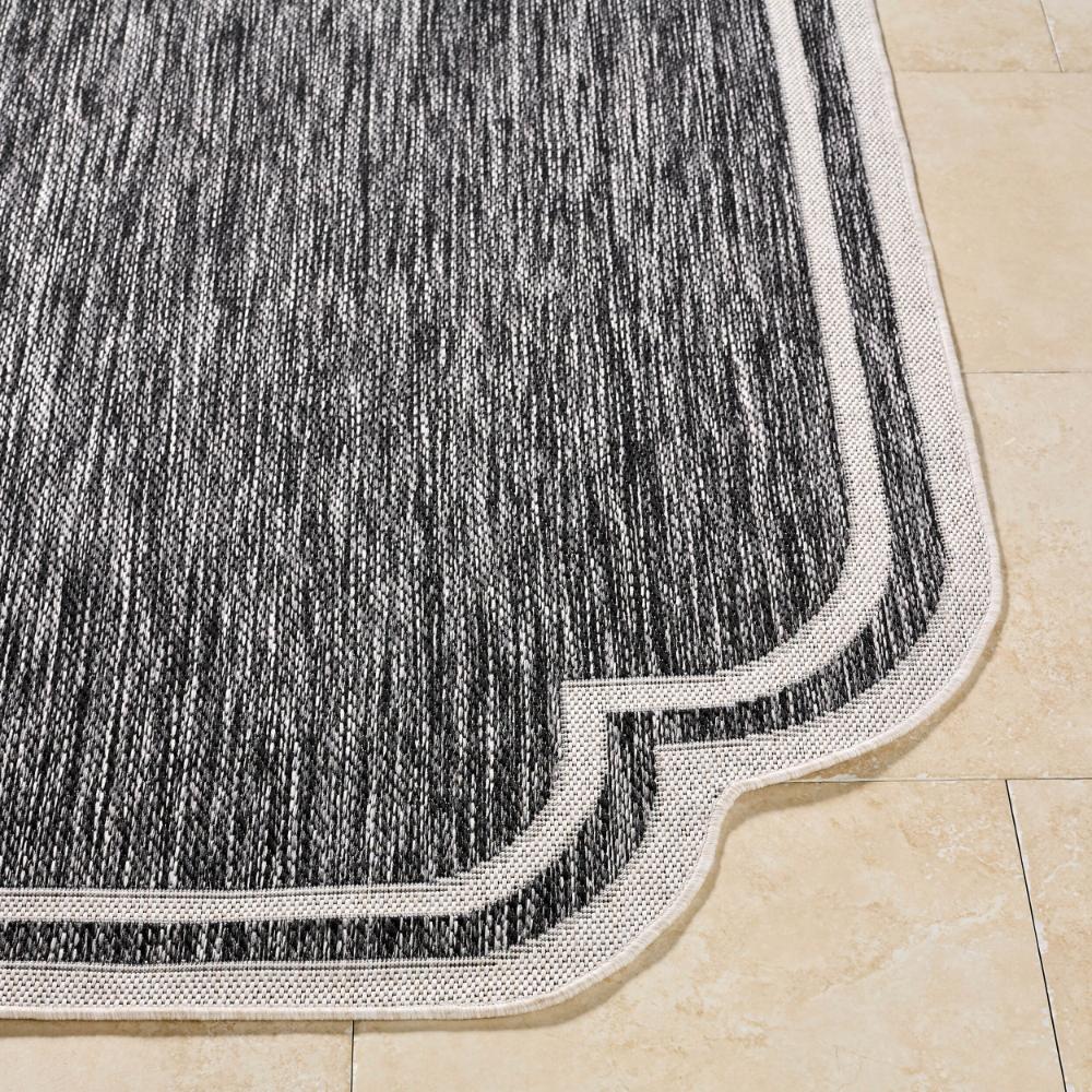 Close-up of the Surya Indoor/Outdoor Scalloped Edge Black + Cream Woven Rug on a beige tiled floor.