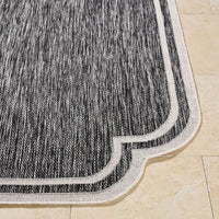 Close-up of the Surya Indoor/Outdoor Scalloped Edge Black + Cream Woven Rug on a beige tiled floor.