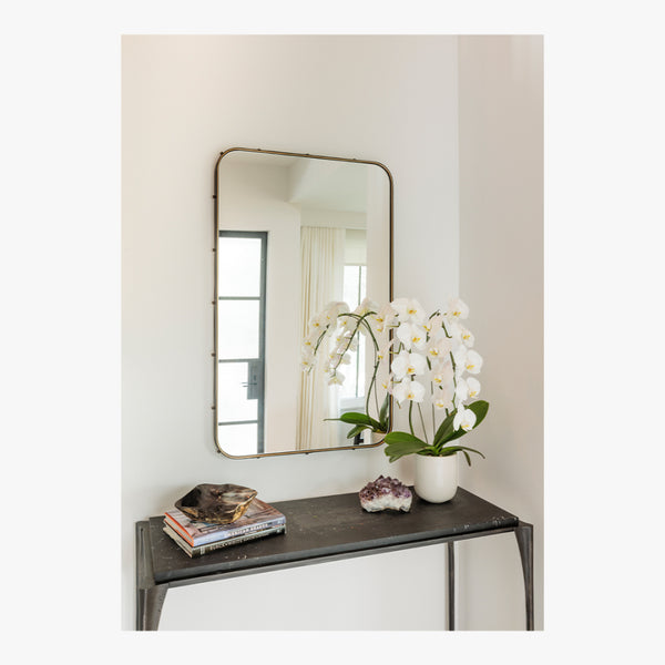 A potted white orchid, books, and crystals on a black table beneath Mirror Home’s Leather Wrapped Antique Bronze Mirror.