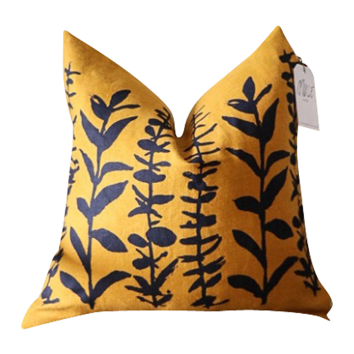 The Mustard + Indigo Eucalyptus Accent Pillow from Muse Textiles is a yellow pillow adorned with a blue leaf pattern and features a white tag attached to the corner. This accent pillow is perfect for adding a touch of color and style to any room.