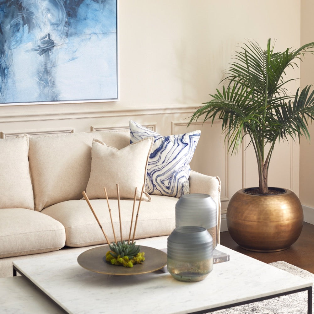The cozy living room is beautifully enhanced by a beige sofa with cushions, a potted palm, and an abstract painting, all complemented by the Cyan Design Nessman Nesting Cocktail Tables featuring white marble tops and a sleek black metal frame.
