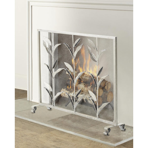 A silver metal fireplace screen with a leaf pattern, shown standing on a glass shelf in front of a white fireplace with a fire burning inside.
