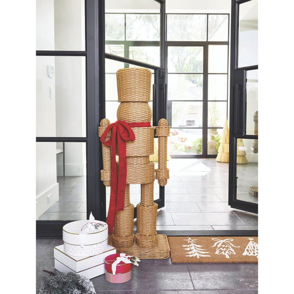 The Santa Barbara Design Studio All Weather Rattan Nutcracker with Red Bow stands by gifts and festive décor near glass doors.
