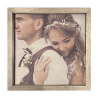 A Thin Wooden Frame Custom Photo Panel by Brayden & Brooks, part of a charming photo gallery wall, features a couple in wedding attire. The woman, wearing a white dress and a flowered hair accessory, hugs the man from behind. The man, dressed in a white shirt and vest, looks away into the distance. Both appear content and serene.