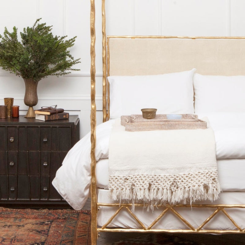 A gold-framed bed with white bedding sits by a black nightstand topped with books, a plant, and Made Goods Opal trays.