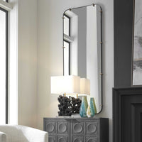 A modern living room corner featuring the Uttermost Pali Dressing Mirror with rounded edges and brushed gold details hanging on a light-colored wall. Below the mirror is a dark, textured cabinet adorned with two blue and green vases and a lamp with a square white shade. A large window is on the left.