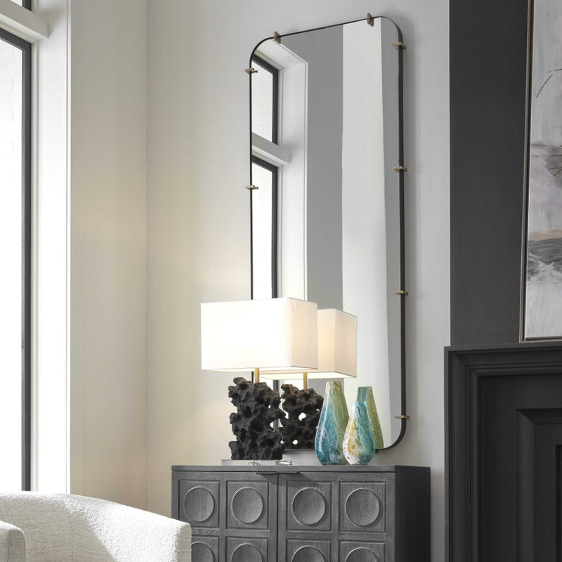 A modern living room corner featuring the Uttermost Pali Dressing Mirror with rounded edges and brushed gold details hanging on a light-colored wall. Below the mirror is a dark, textured cabinet adorned with two blue and green vases and a lamp with a square white shade. A large window is on the left.
