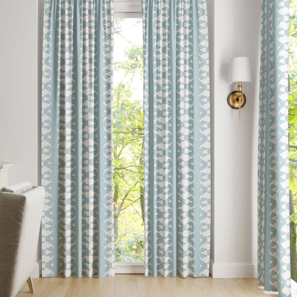 Laura Park Designs' Palm Mist Curtain Panels in light blue with a white geometric pattern hang by a window.