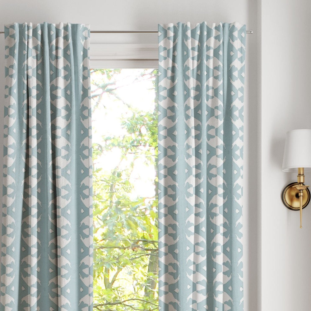 Palm Mist Curtain Panels by Laura Park Designs frame a window with greenery, creating a calm look; wall sconce on right.
