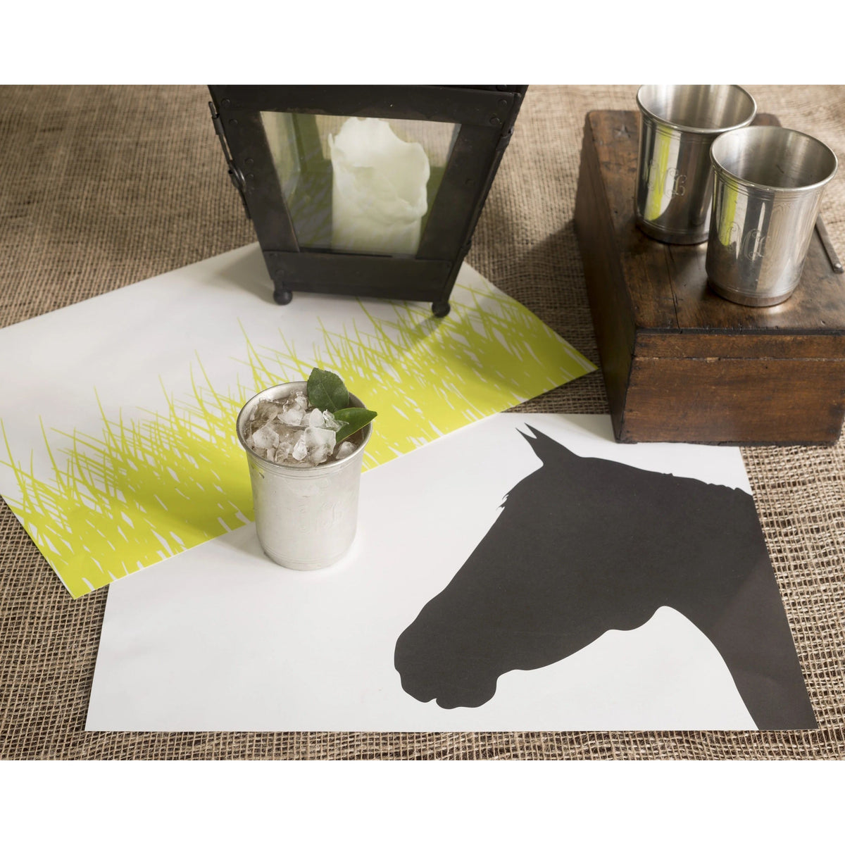 A metal cup with a drink sits on a Plat du Jour Spring Grass Paper Placemat Pad, adding charm to the table.
