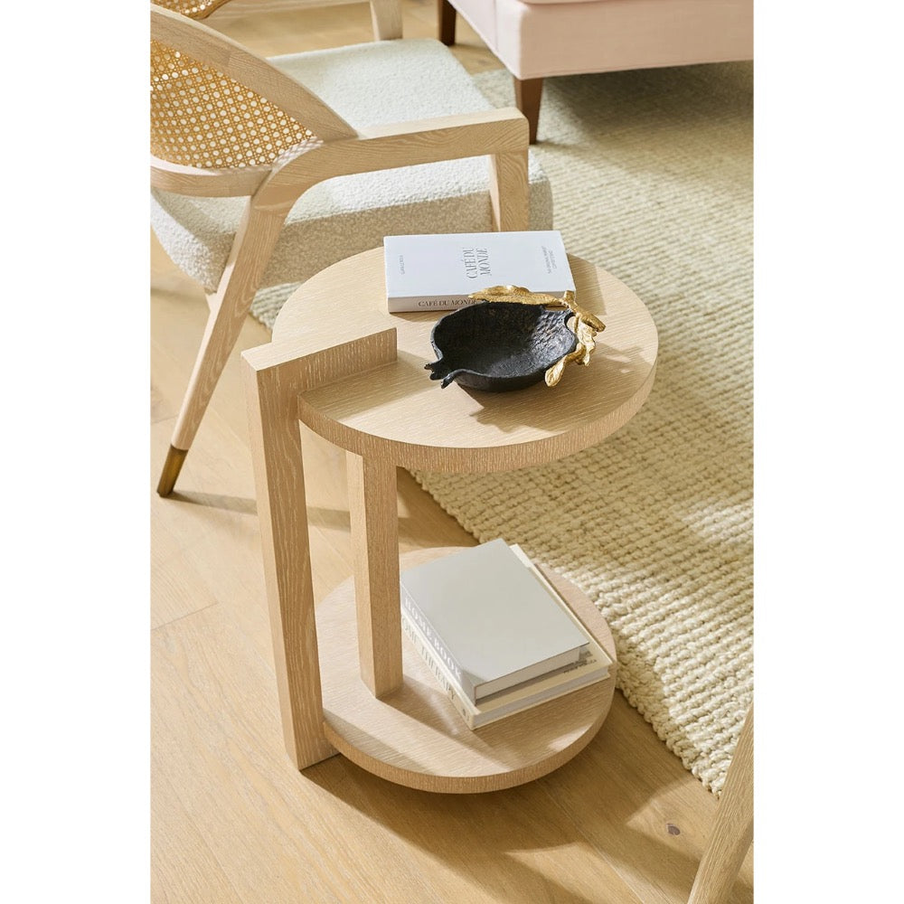 A light natural wood side table with two circular tiers, styled with books and a small black bowl in a bright, modern room setting.