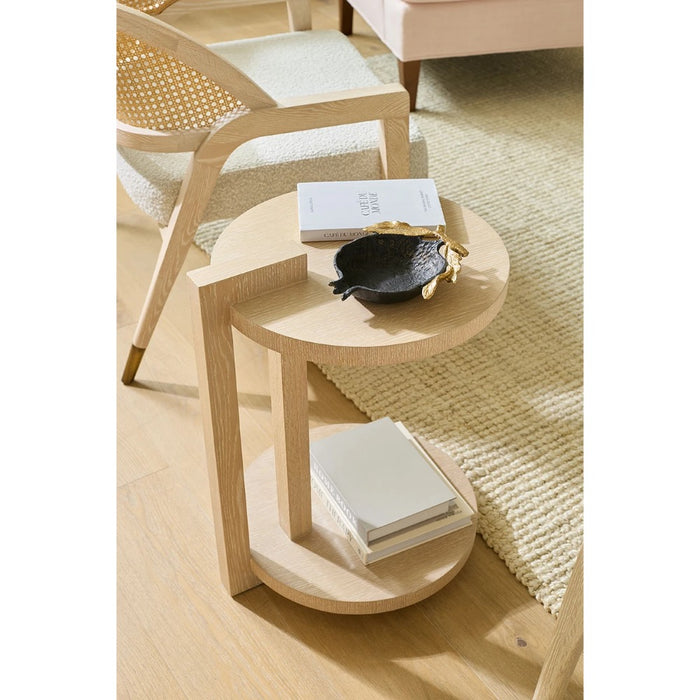 A light natural wood side table with two circular tiers, styled with books and a small black bowl in a bright, modern room setting.