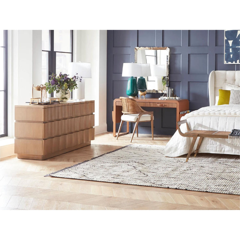 Modern bedroom with wooden dresser, vanity desk with green lamps, chair, upholstered bed, and checkered rug on hardwood floor.