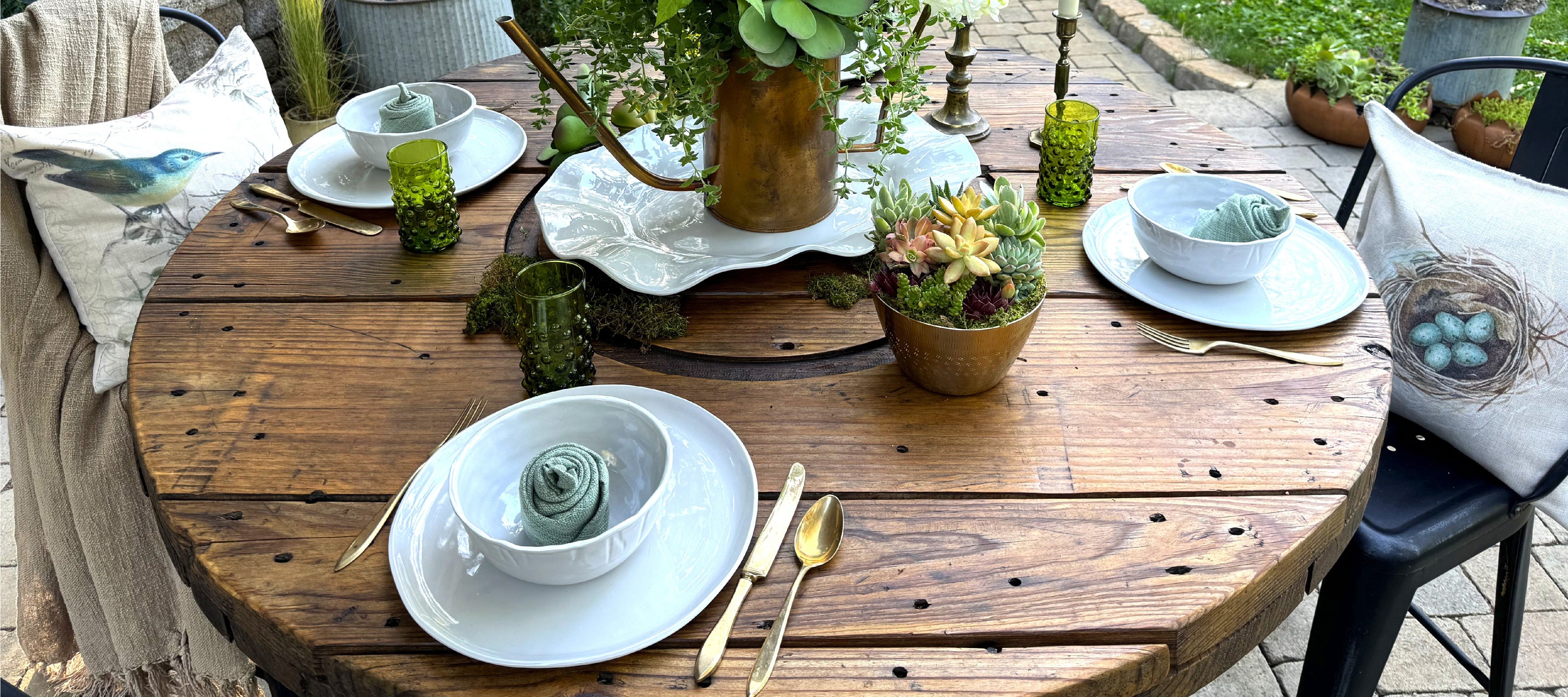 A rustic outdoor table set for four, with white plates, gold cutlery, green glasses, and potted plants as a centerpiece.