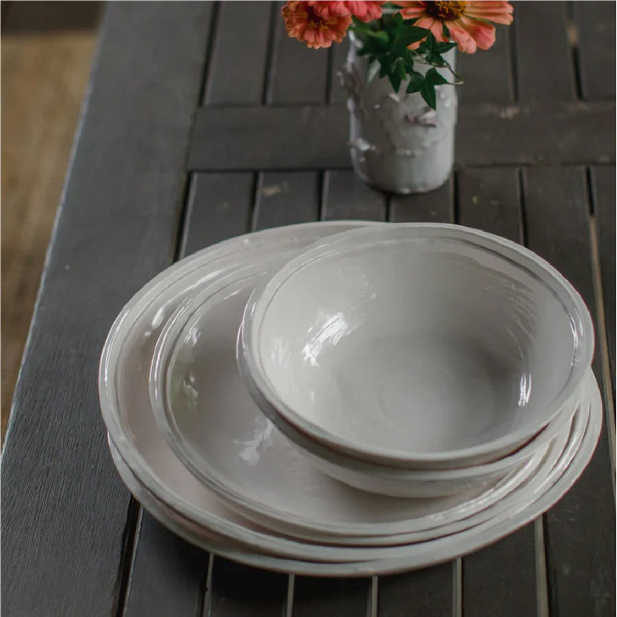 A stack of light-colored Double Line Stone Melamine Dinner Plates by Always Relish sits on a wooden table, accompanied by a small vase of flowers. The setup invites mixing and matching, ensuring a charming dining experience.