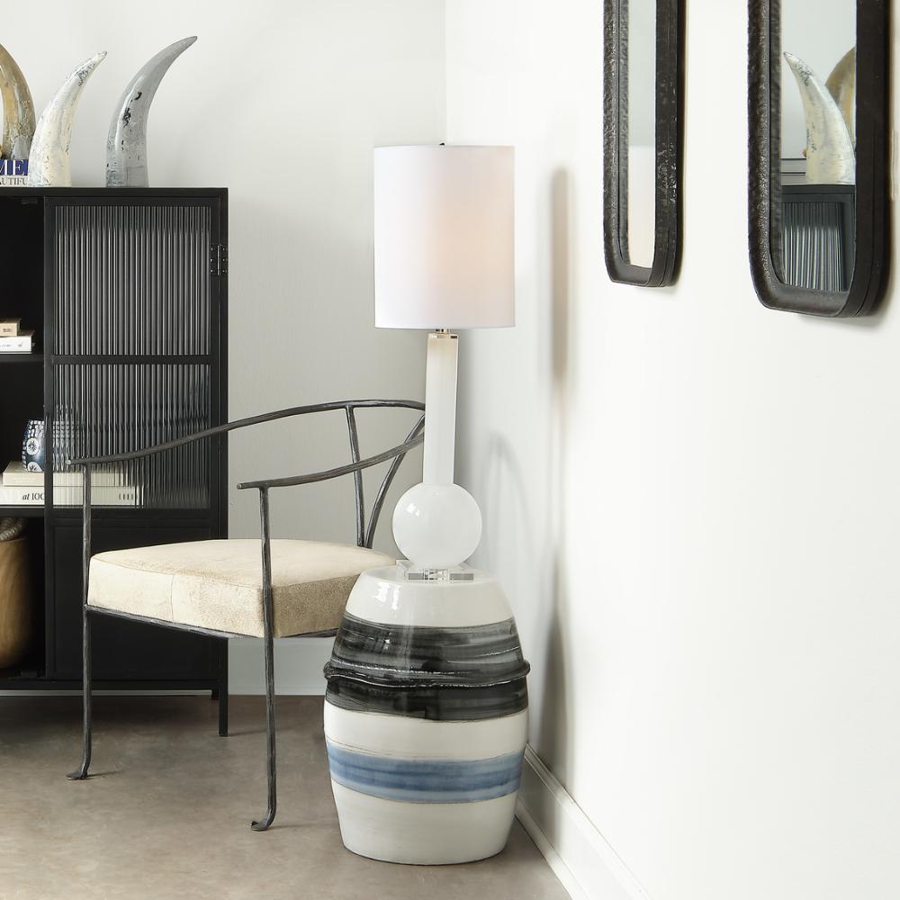 A modern table lamp with a white drum shade, a white cylindrical stem, and a glossy ceramic side table featuring black and blue horizontal stripes, styled in a contemporary room next to a black metal chair.