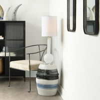 A modern table lamp with a white drum shade, a white cylindrical stem, and a glossy ceramic side table featuring black and blue horizontal stripes, styled in a contemporary room next to a black metal chair.