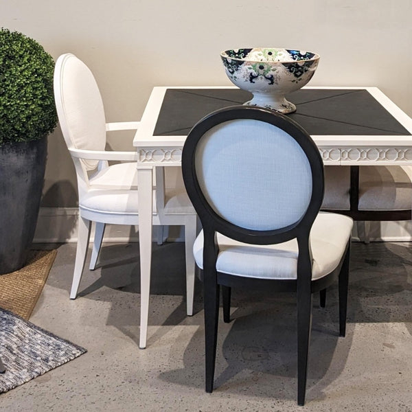 A dining room scene with a white table and upholstered chairs. The foreground chair has a black frame and light blue fabric upholstery, while the left chair is white.