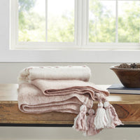 The Blush Rose + Ivory Plush Bell Mohair Throw by India's Heritage rests folded on a bench by a window with greenery.