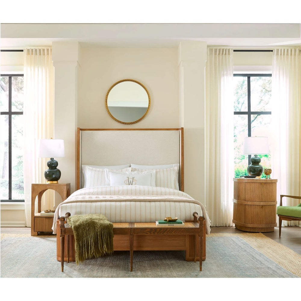 A mid-century modern wooden bed frame with a tall upholstered headboard, styled in a bright, neutral-toned bedroom with cream curtains, a wooden bench, and matching nightstands.