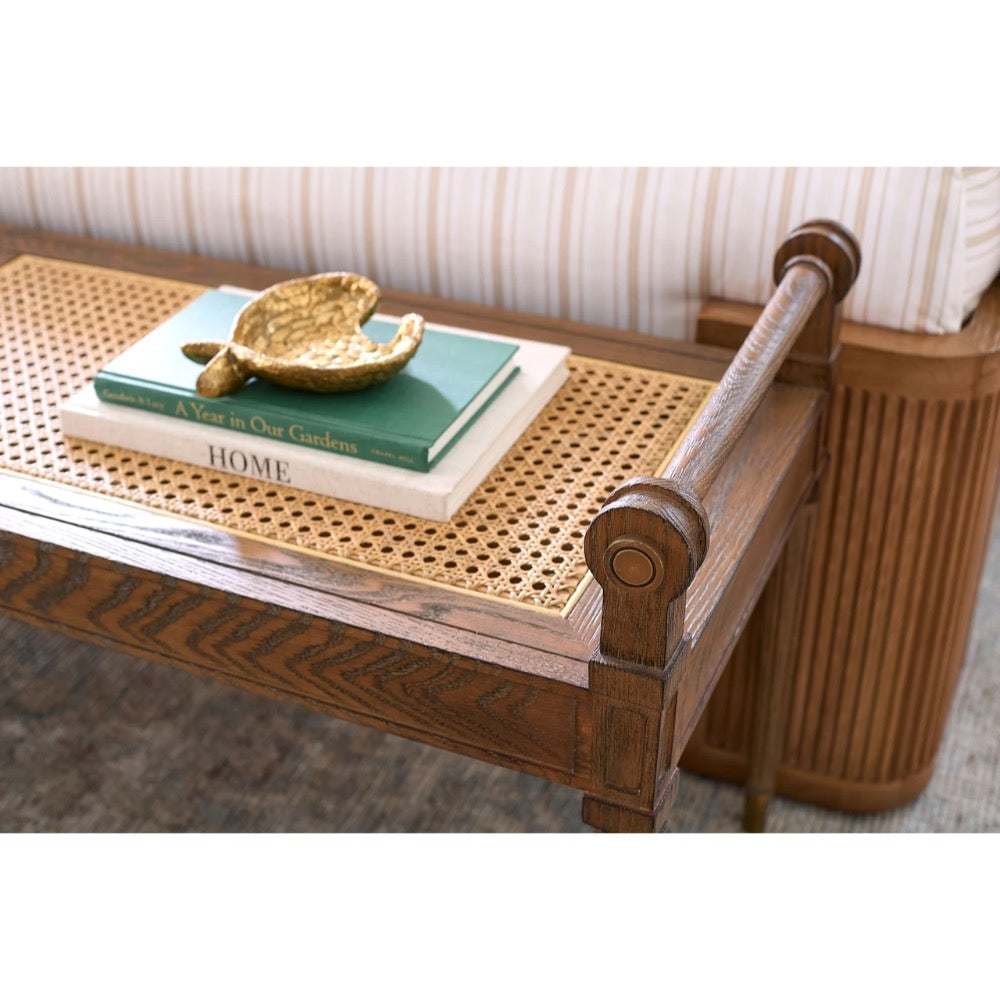A close-up of a wooden bench or ottoman with a woven cane top, styled with stacked books and a gold decorative object, in front of a striped cushion.