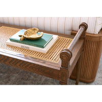 A close-up of a wooden bench or ottoman with a woven cane top, styled with stacked books and a gold decorative object, in front of a striped cushion.