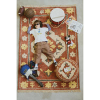 A child lies on a large patterned area rug surrounded by playful props, including a football, basketball, drum, guitar, round pouf, and small stuffed toy, photographed from above on a light floor.