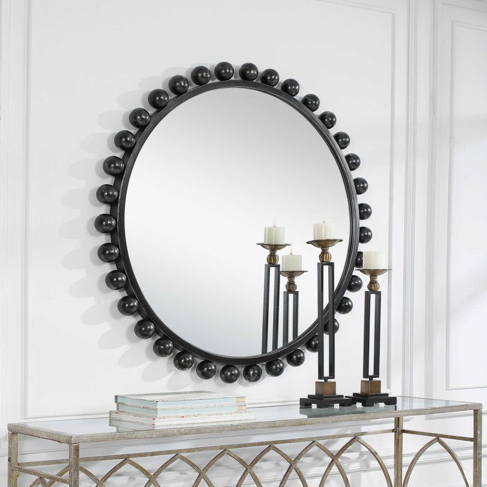 The Cyra Burst Black Mirror by Uttermost becomes the focal point above a console table adorned with books and candle holders in a crisp white room.