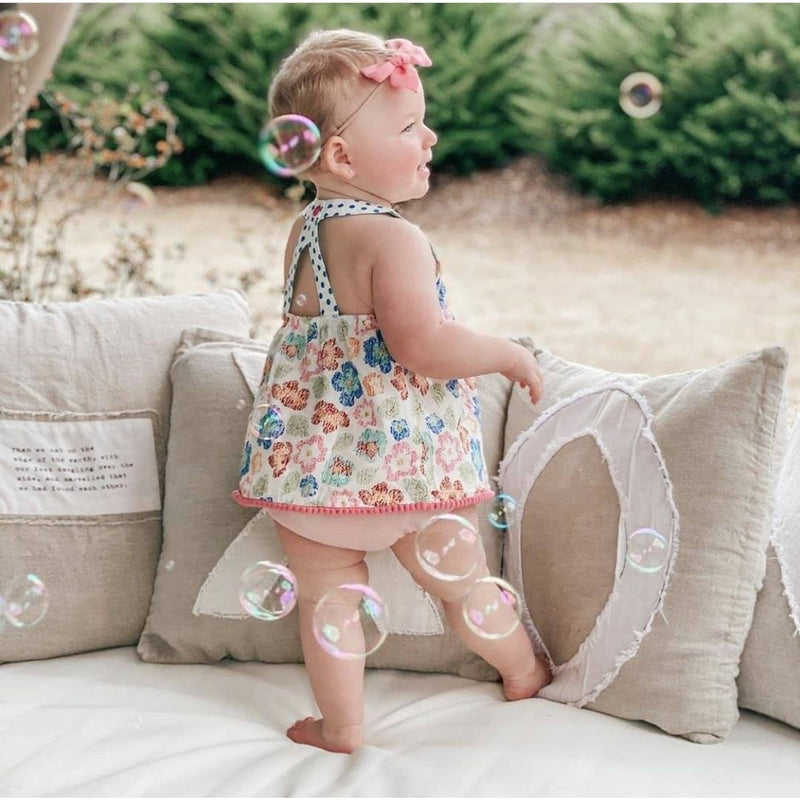 A baby in a floral dress stands on a couch surrounded by bubbles, creating a vintage vibe with Sugarboo & Co.'s stonewashed pillow.