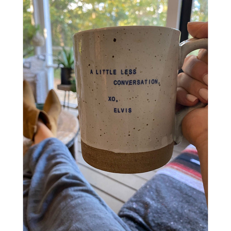 A person cradles a whimsical Sugarboo & Co. XO Legends Coffee Mug (1st Edition) from the set of 8, adorned with the words "A little less conversation, XO, Elvis," while unwinding on a cozy, covered porch.