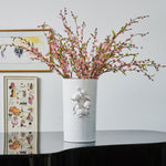 The Pomegranate Vase by Abigails, with its intricate designs, holding pink blossoms on top of a glossy black piano, framed artworks in the background, exudes a modern aesthetic.