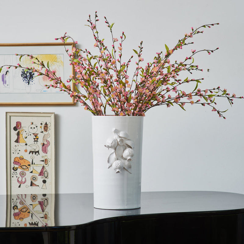 The Pomegranate Vase by Abigails, with its intricate designs, holding pink blossoms on top of a glossy black piano, framed artworks in the background, exudes a modern aesthetic.